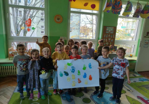 Dzieci trzymają laurkę zrobioną dla kolegi w tle trzy okna i zielone ściany