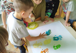 dzieci stoją przy stole i odbijają w farbie ręce na kartonie tworzą prace plastyczną