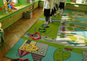 Dzieci stoją na kolorowym dywanie na którym ułożone są kolorowe szarfy