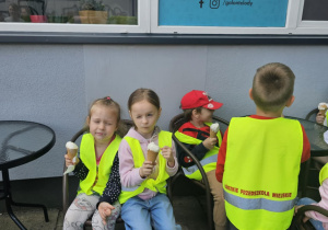 Dzieci siedzą przed lodziarnią i jedzą lody.