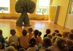 Dzieci siedzą na dywanie oglądają występ cyrkowy, na środku sali znajduje się artysta w zabawnym, kolorowym kostiumie przypominającym spręzynę lub tunel.W tle widoczne okna z kolorowymi roletami.