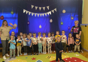 Na zdjęciu widać dzieci stojące w rzędzie tło niebieskie z kolorowym napisem ,,DZIEŃ PRZEDSZKOLAKA''