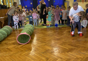 Na zdjęciu widać hol przedszkolny dzieci biorą udział w konkurencjach ruchowych na pierwszym planie ustawione są kolorowe tunele. Z boku widać dziewczynkę chodzacą na specjonych ,,kubeczkowych szczudłach'' na sznurkach obok stoją panie nauczycielki które czuwają nad przebiegiem zabawy