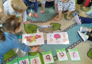 Dzieci siedzą na kolorowym dywanie oglądają litery i obrazki związane z jabłkiem