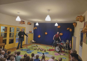 Mężczyzna gra na instrumentach - perkusji i bębnach, dzieci obserwują go.