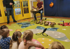 Mężczyzna gra na instrumentach - perkusji i bębnach, dzieci obserwują go.
