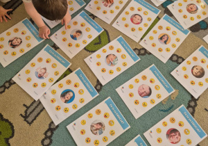 Dzieci siedzą na dywanie i oglądają zdjęcia twarzy innych dzieci przedstawiających różne emocje. Dzieci wskazują emocje o które prosi pani.