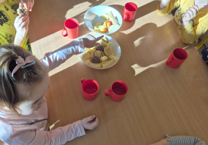 Dzieci siedzą przy stole. Dzieci jedzą słodkie ciasta, jedzą owoce, jabłka, piją sok.