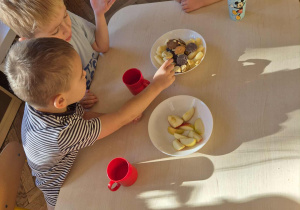Dzieci siedzą przy stole, jedzą słodkie ciastka, piją sok z kolorowych kubeczków.