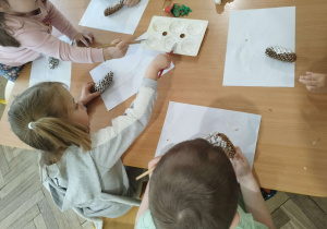 Dzieci siedzą przy stolikach wykonują prace plastyczną malują szyszkę białą farbą