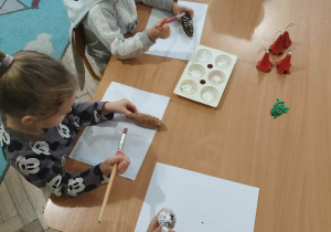 Dzieci siedzą przy stolikach wykonują prace plastyczną malują szyszkę białą farbą