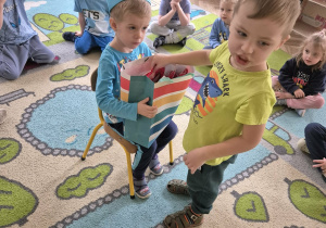 Jeden z chłopców siedzi i na krześle z koroną na głowie i trzyma torebkę. Drugi z chłopców wyciąga z torebki prezent i składa mu życzenia.