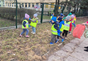 Pani trzyma worek na śmieci. Dzieci rzucają do worka zebrane śmieci z trawy.