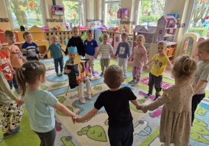 Dzieci stoją w kole i trzymają się za ręce. Dziewczynka siedzi na krześle w środku koła i trzyma prezenty.