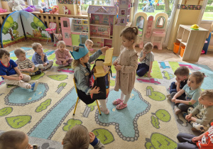 Dzieci siedzą w kole. Dziewczynka składa życzenia dziewczynce, która ma urodziny.