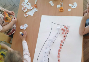 Dzieci biorą udział w zajęciach plastycznych. Na środku stołu leży duży arkusz papieru z narysowanym kształtem flagi. Dzieci wspólnie go ozdabiają.